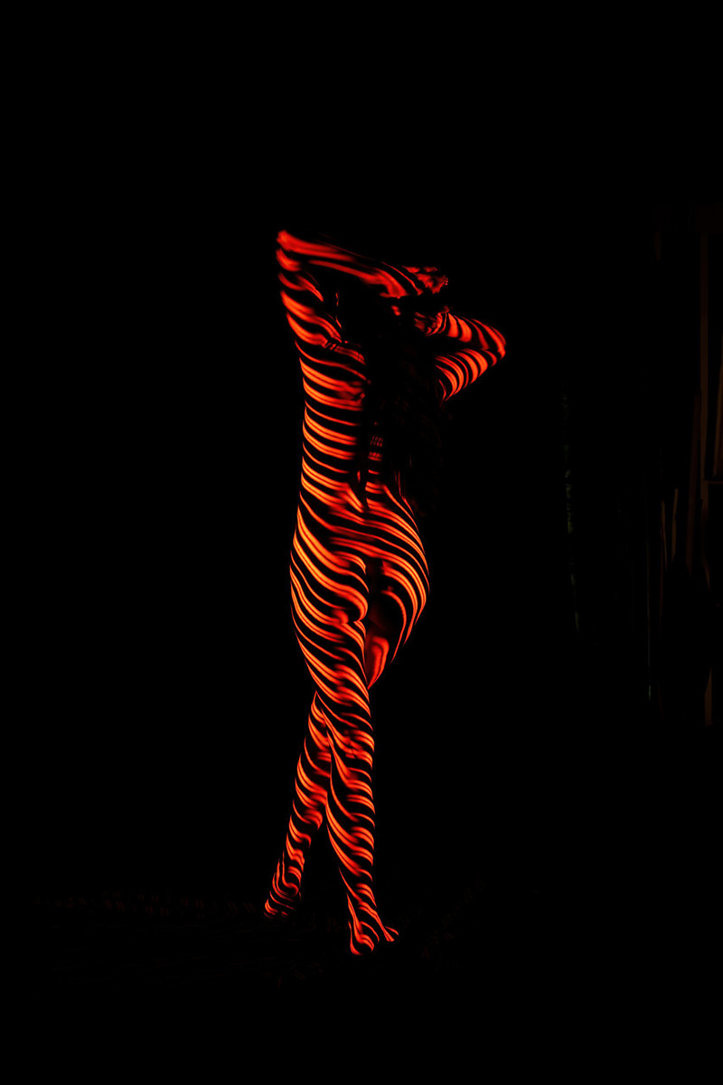 Nude person standing with arms raised, covered in red and black striped light patterns against a dark background