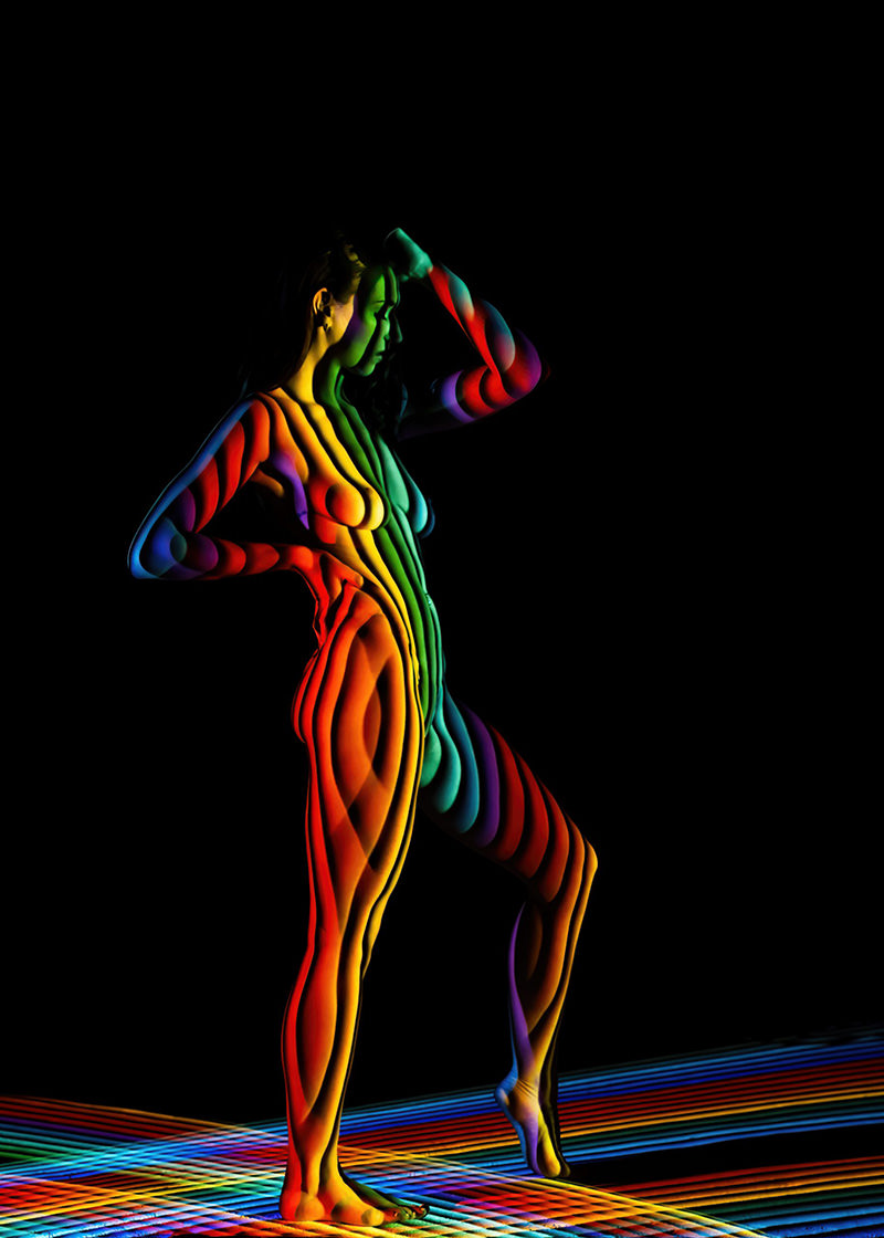 A nude human figure striking a pose against a black background, covered in vivid rainbow-colored light stripes that wrap around the body and extend onto the floor