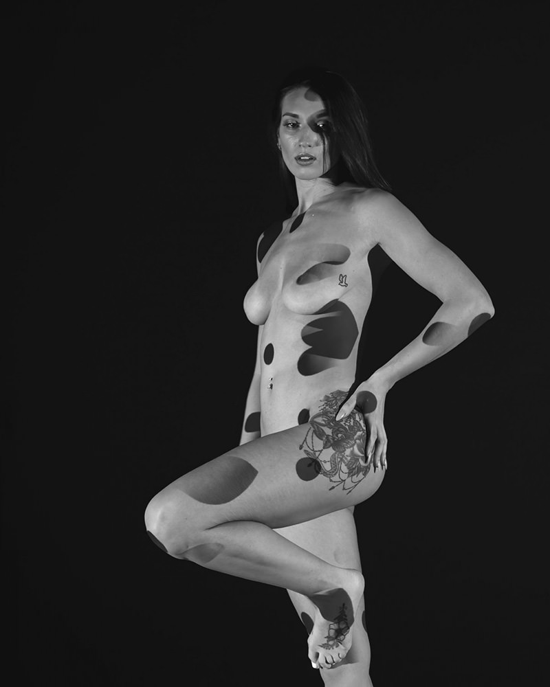 Nude woman standing on one leg against a black background, with abstract circular shadows projected on her body and visible tattoos on her thigh and foot