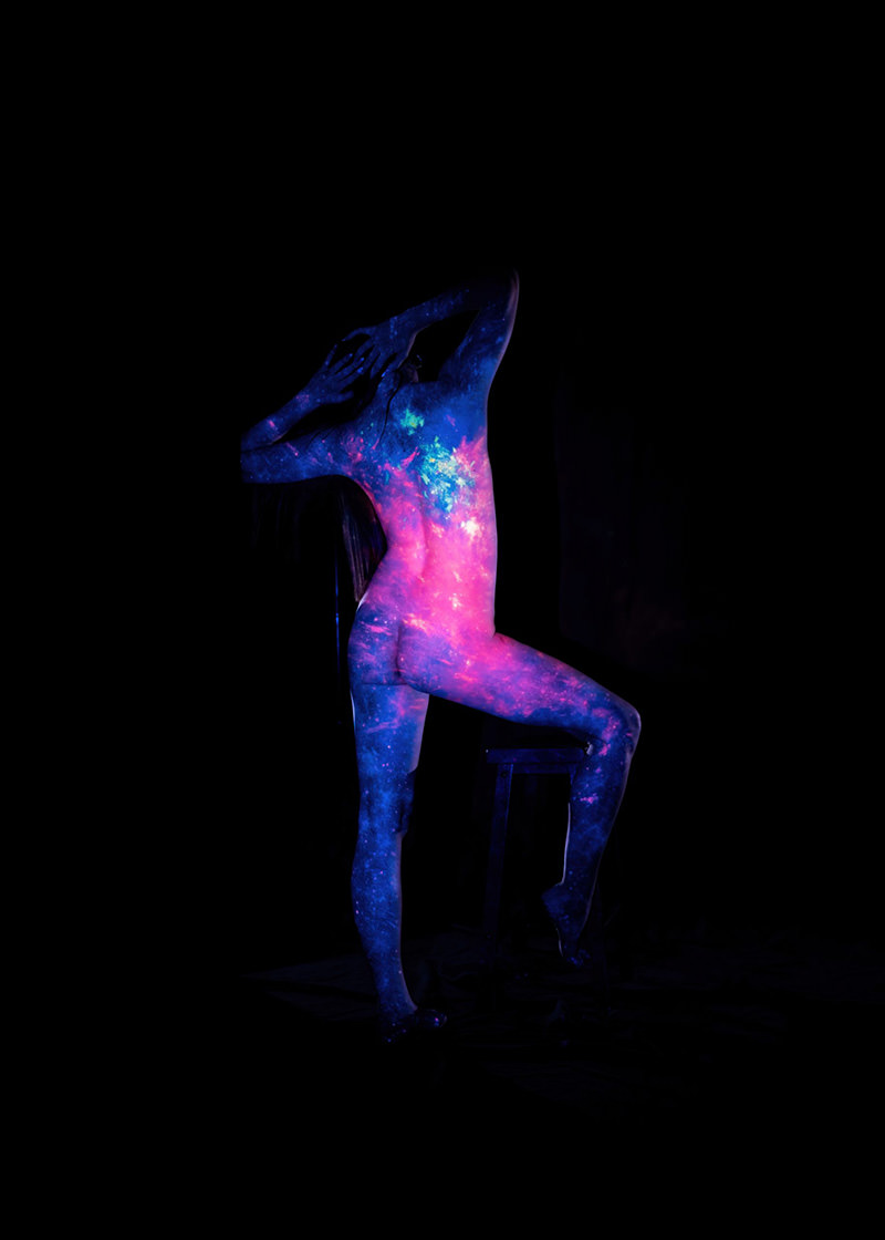 Nude person standing on one leg with arms raised, body painted with vibrant cosmic and galaxy patterns glowing in UV light against a black background