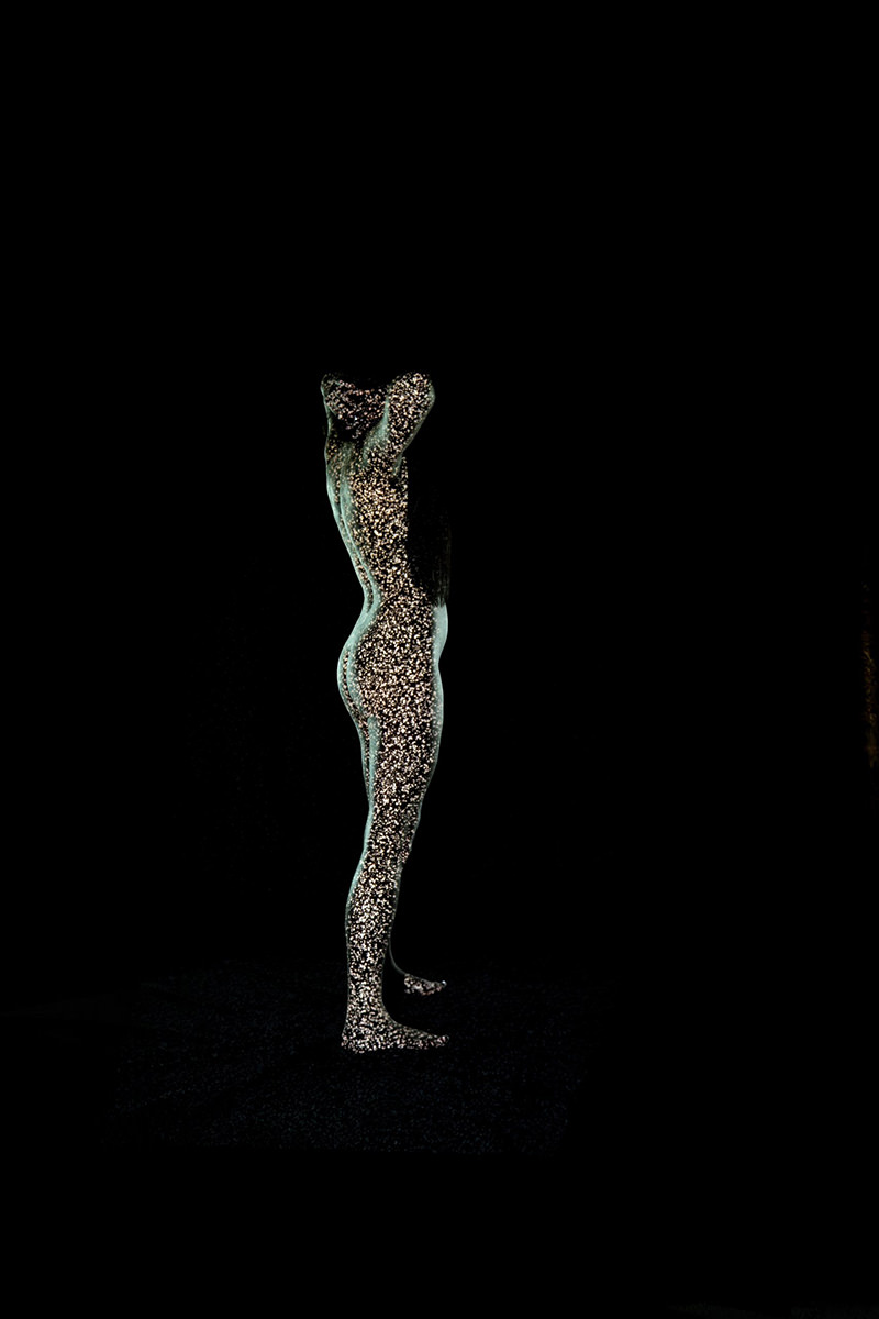 Side view of a nude human figure covered in sparkling lights, standing against a black background with arms raised behind the head