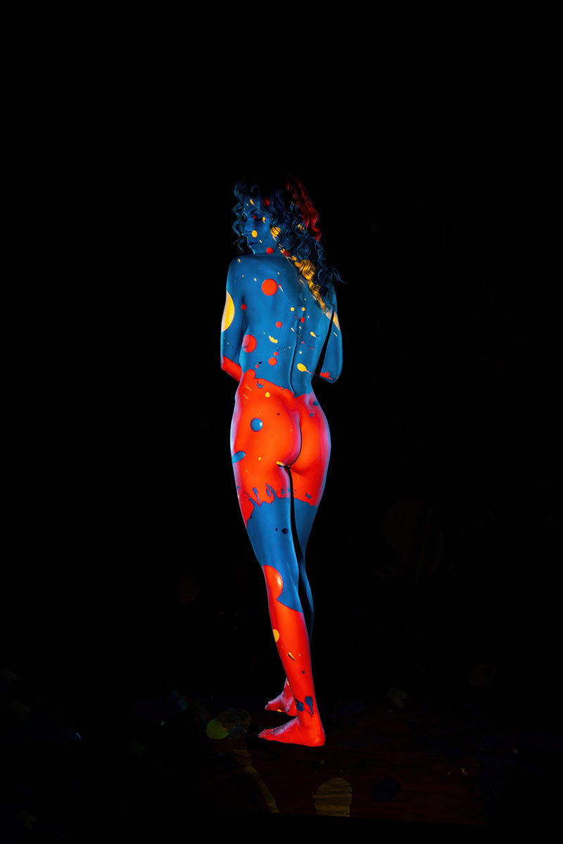 Nude person standing with back to the camera, body painted in bright blue and red with yellow and orange dots and splashes, against a black background