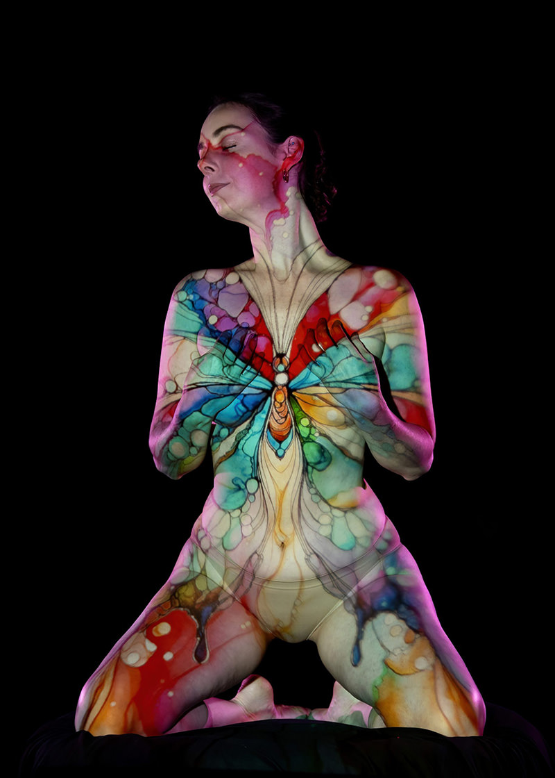Person kneeling with eyes closed, colorful butterfly design projected on their upper body and face against black background