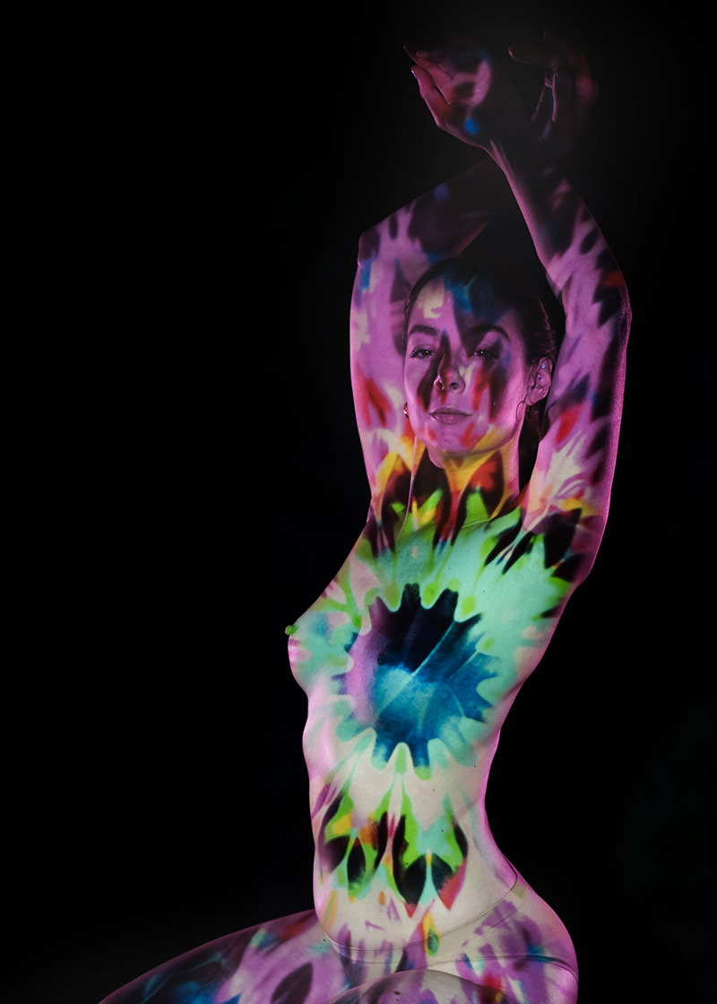 Woman with arms raised above head, illuminated by vibrant tie-dye pattern projection on her skin, set against a black background