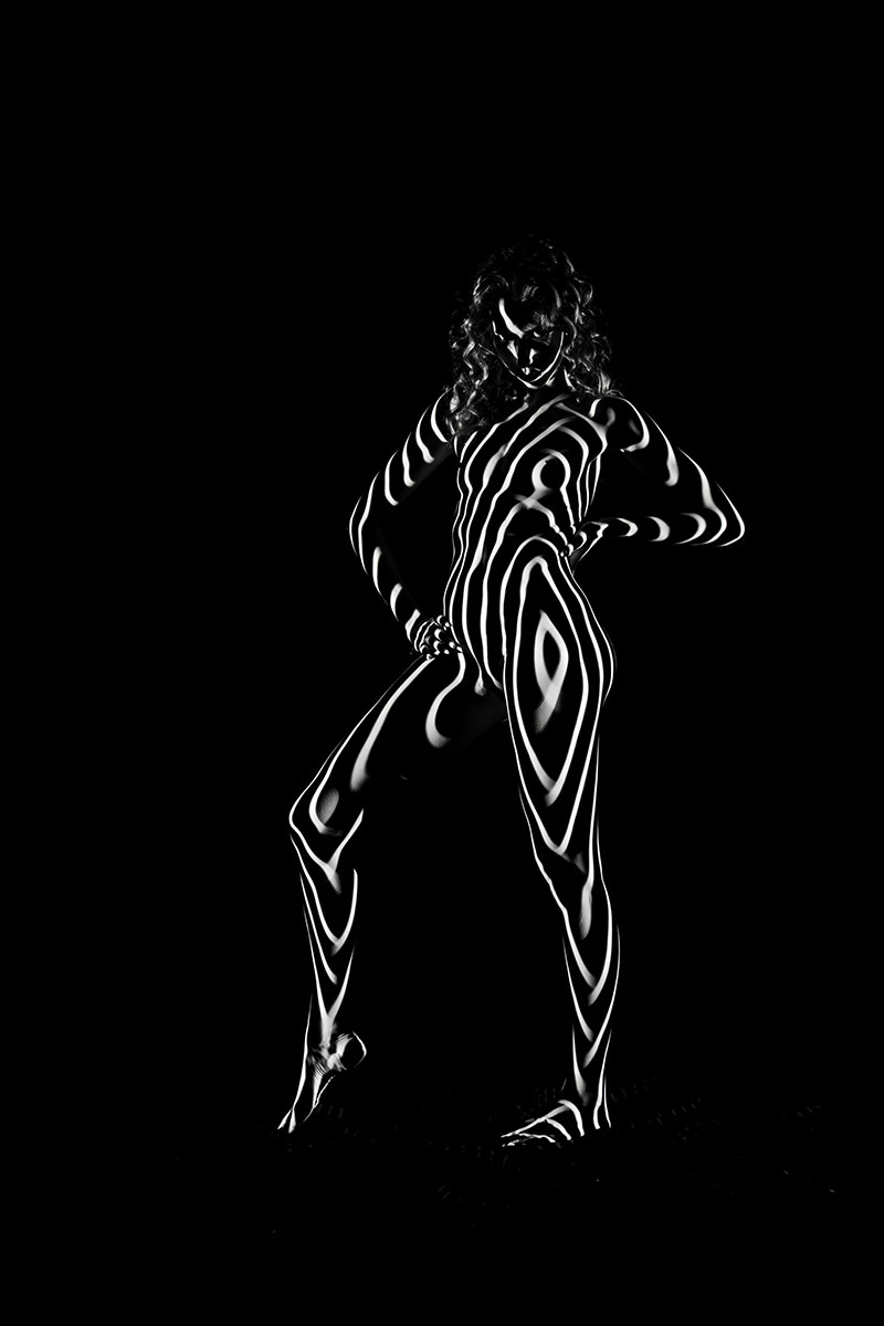 Nude person standing with hands on hips, covered in bright white spiral and curved stripes that contrast sharply against a black background