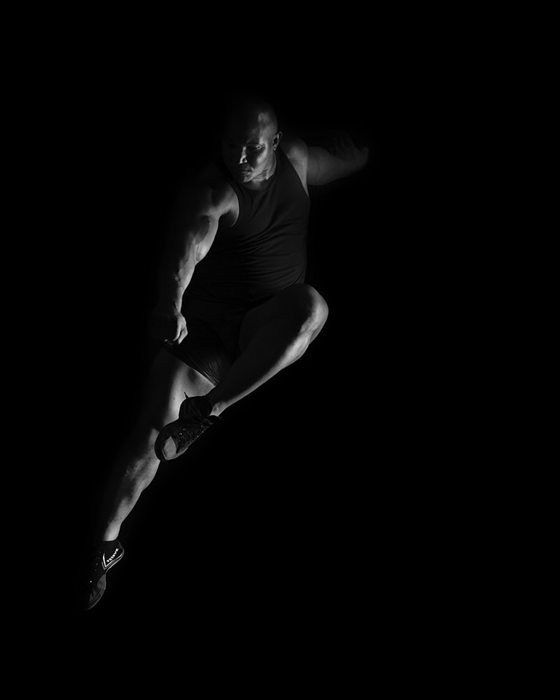 Muscular athlete performing a dynamic jump against a black background, wearing a sleeveless top and athletic shoes