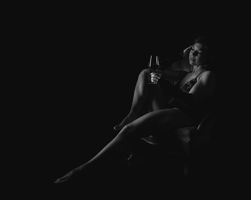 Woman seated on an armchair wearing fishnet stockings and lace lingerie, holding a glass of wine, with soft light highlighting her face and legs against a dark background