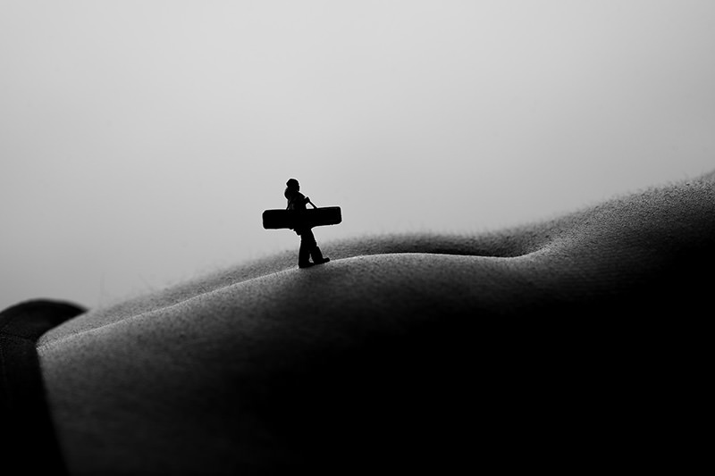 Miniature silhouette figure of a person carrying a surfboard walking on a textured, curvy surface resembling sand dunes or waves in black and white
