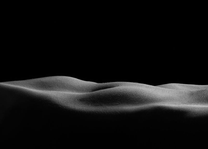 Close-up black and white image showing smooth curves and texture of a human body in a reclining position, highlighting the natural shapes and shadows resembling gentle hills.