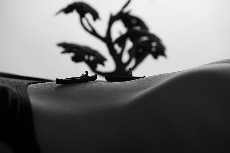 Two small canoes with figurines floating on a curved surface resembling a human body, with a blurred tree in the background