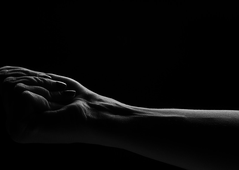 Close-up of two hands clasped together with visible veins and textures, lit dramatically against a black background