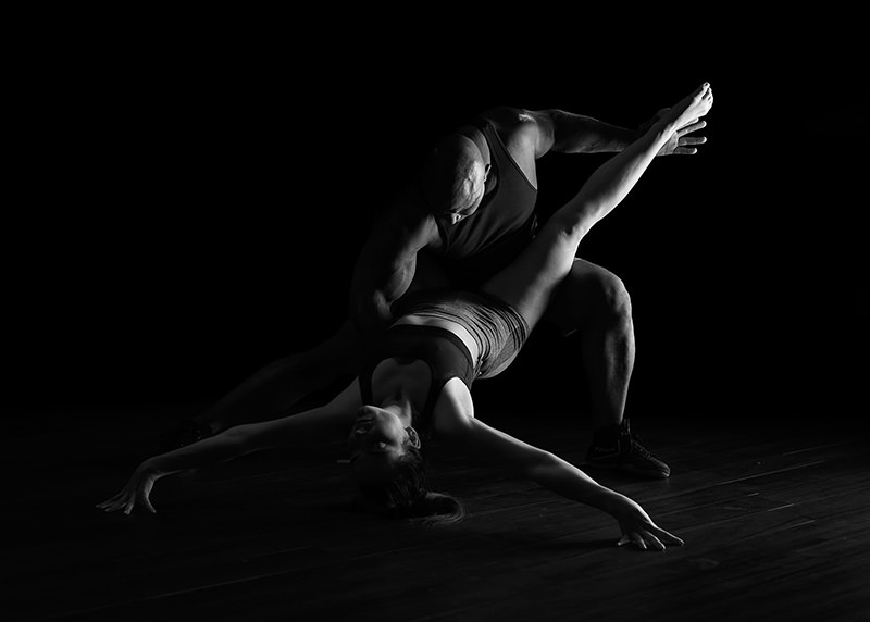 A muscular man supports a woman in a dramatic dance pose; she arches backward with one leg extended upward and arms reaching out, set against a dark background.