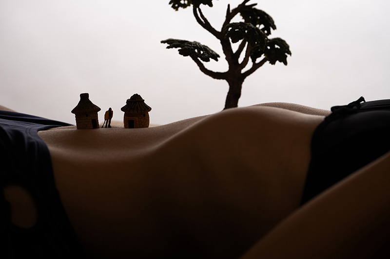 Curved torso of a person wearing dark underwear with two miniature huts, a small figure, and a tree arranged on the skin resembling a village scene