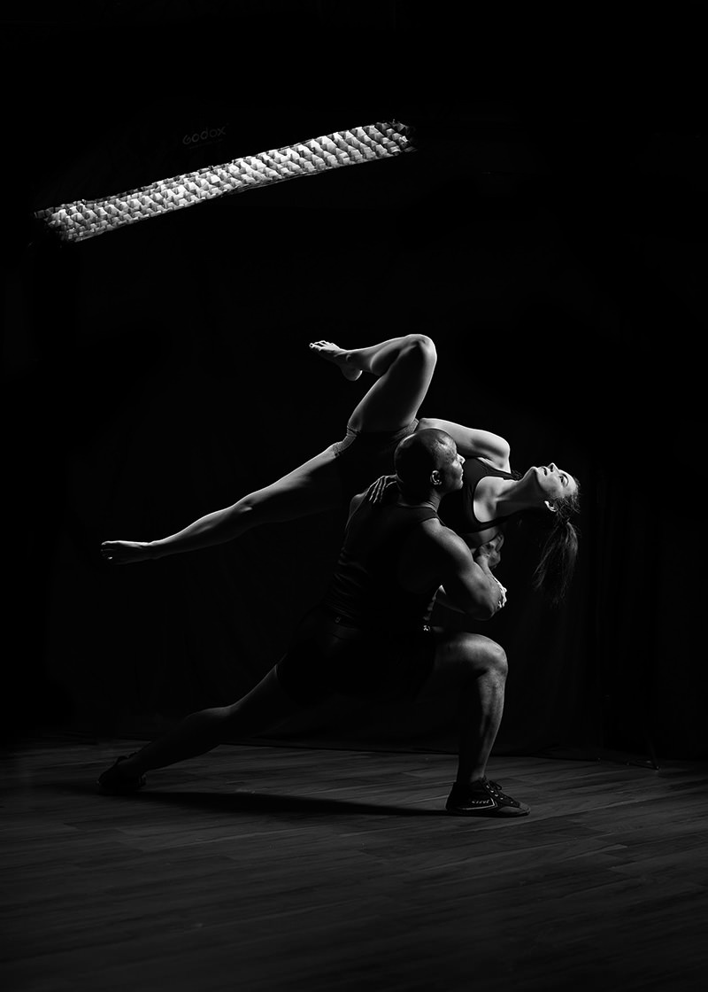 Muscular man supporting a woman in a dance pose with one leg extended, both illuminated against a dark background with a softbox light overhead