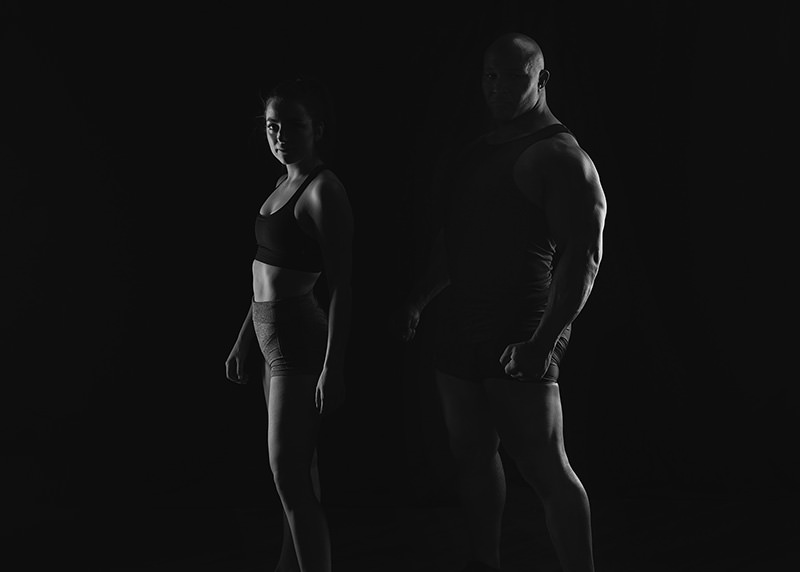 A fit woman and muscular man standing side by side against a black background, highlighted by dramatic low-key lighting that emphasizes their athletic physiques and contrasts their body shapes.
