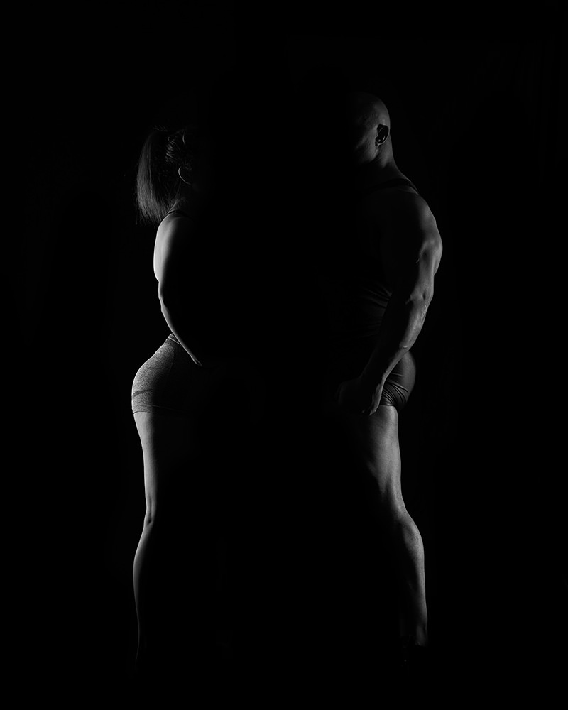 Side profiles of a muscular man and woman standing back to back, dimly lit against a black background highlighting their toned bodies