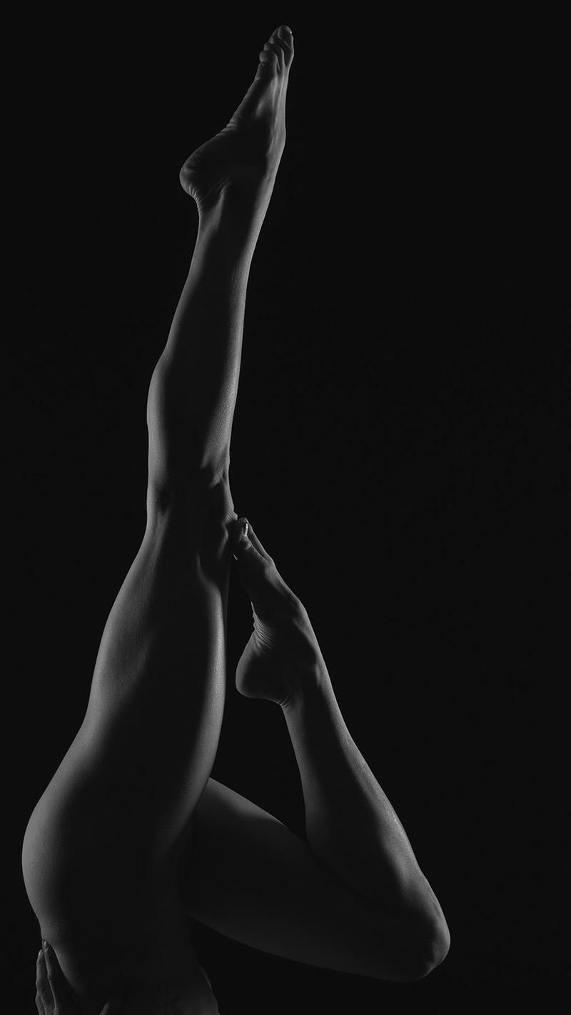 close-up of a person’s legs in a vertical pose with one leg extended straight up and the other bent, set against a black background with soft lighting highlighting muscle contours