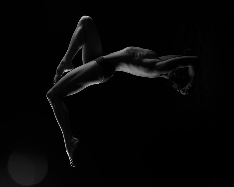 A person performing a backbend bridge pose, their body illuminated against a black background, highlighting muscular definition and a tattoo on the side.