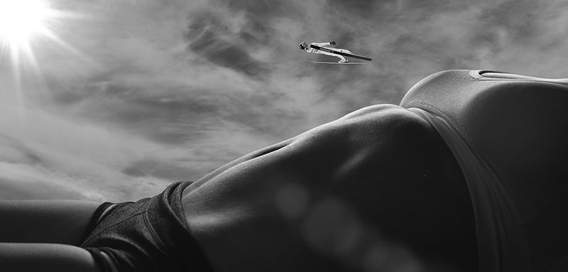 Low-angle view of a person lying down wearing a sports bra and shorts with a ski jumper captured mid-air against a cloudy sky and bright sun