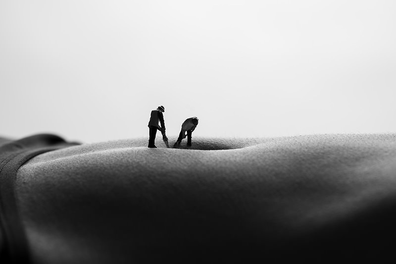 Two tiny figurines of workers positioned on a close-up of a human torso, appearing to dig into the skin near the navel in black and white