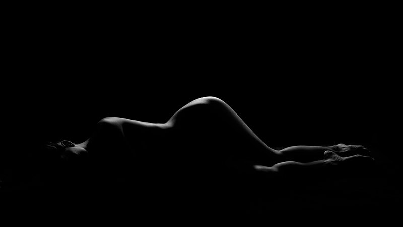 Nude person lying on their side with soft lighting highlighting the curved contours of their body against a dark background