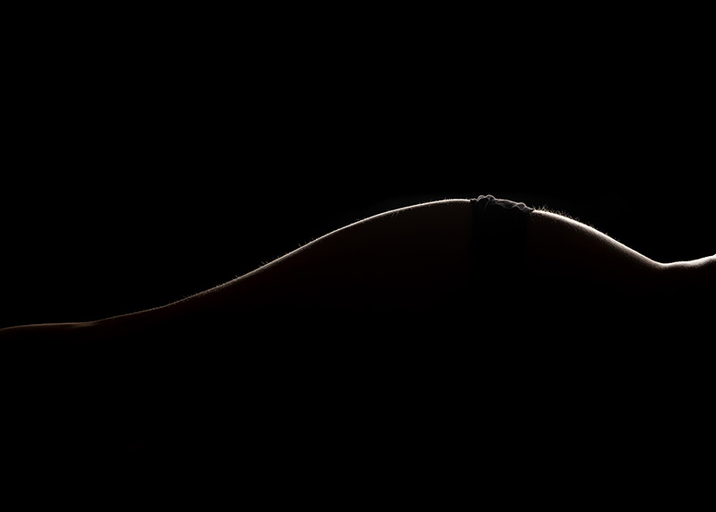 Close-up side view of a person's reclining body with soft lighting outlining the curves against a black background