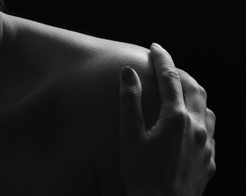 Close-up of a hand gently touching a bare shoulder against a dark background with soft lighting highlighting the skin texture