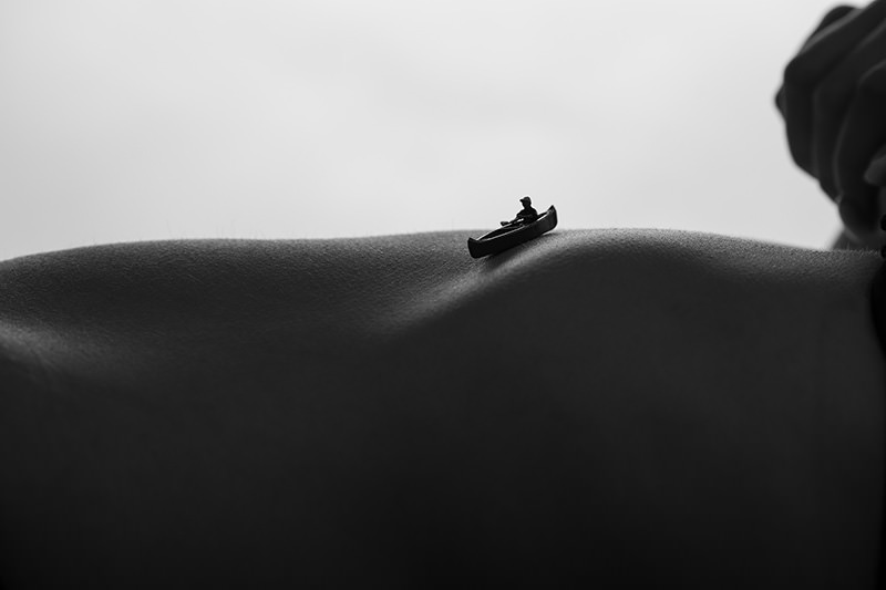 A tiny figure in a canoe positioned on a curving, textured surface resembling a natural landscape, all in black and white.