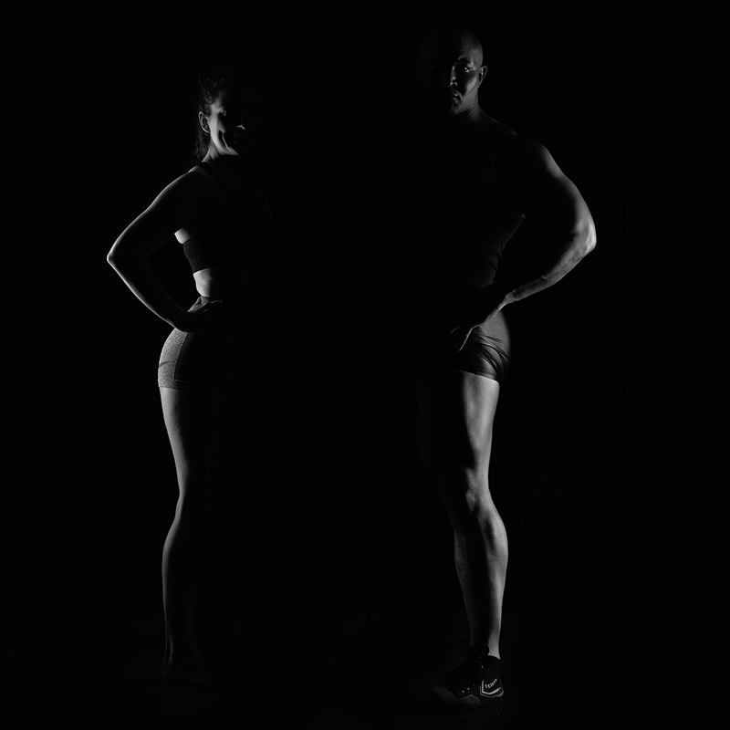 Man and woman standing side by side wearing athletic shorts and tops, their bodies partially illuminated against a dark background, showing muscular definition and confident poses.