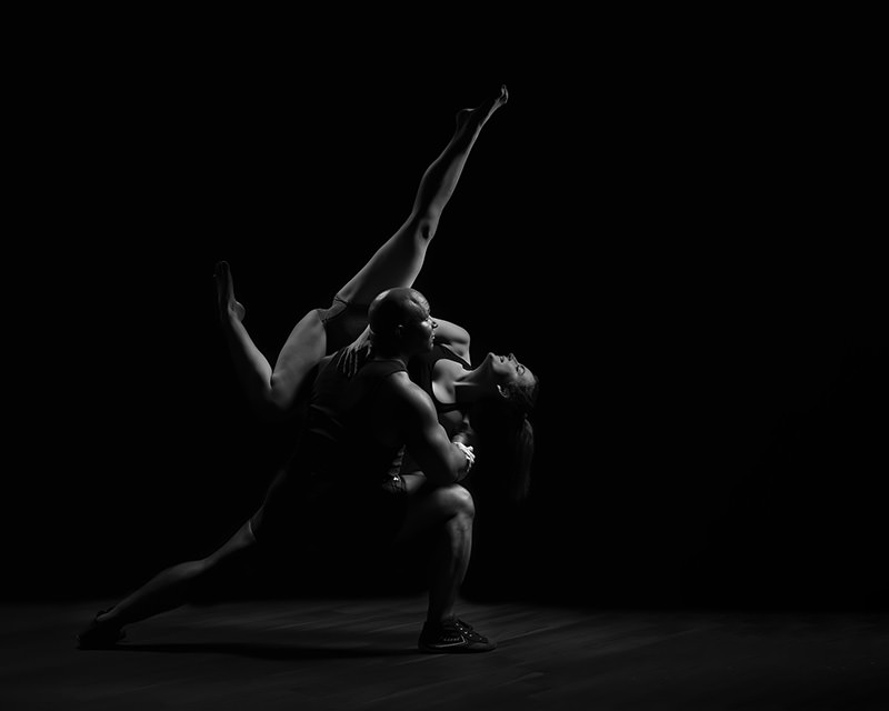 A male and female dancer perform an intricate pose; the man supports the woman who extends one leg high in a dramatic, balanced stretch against a dark background