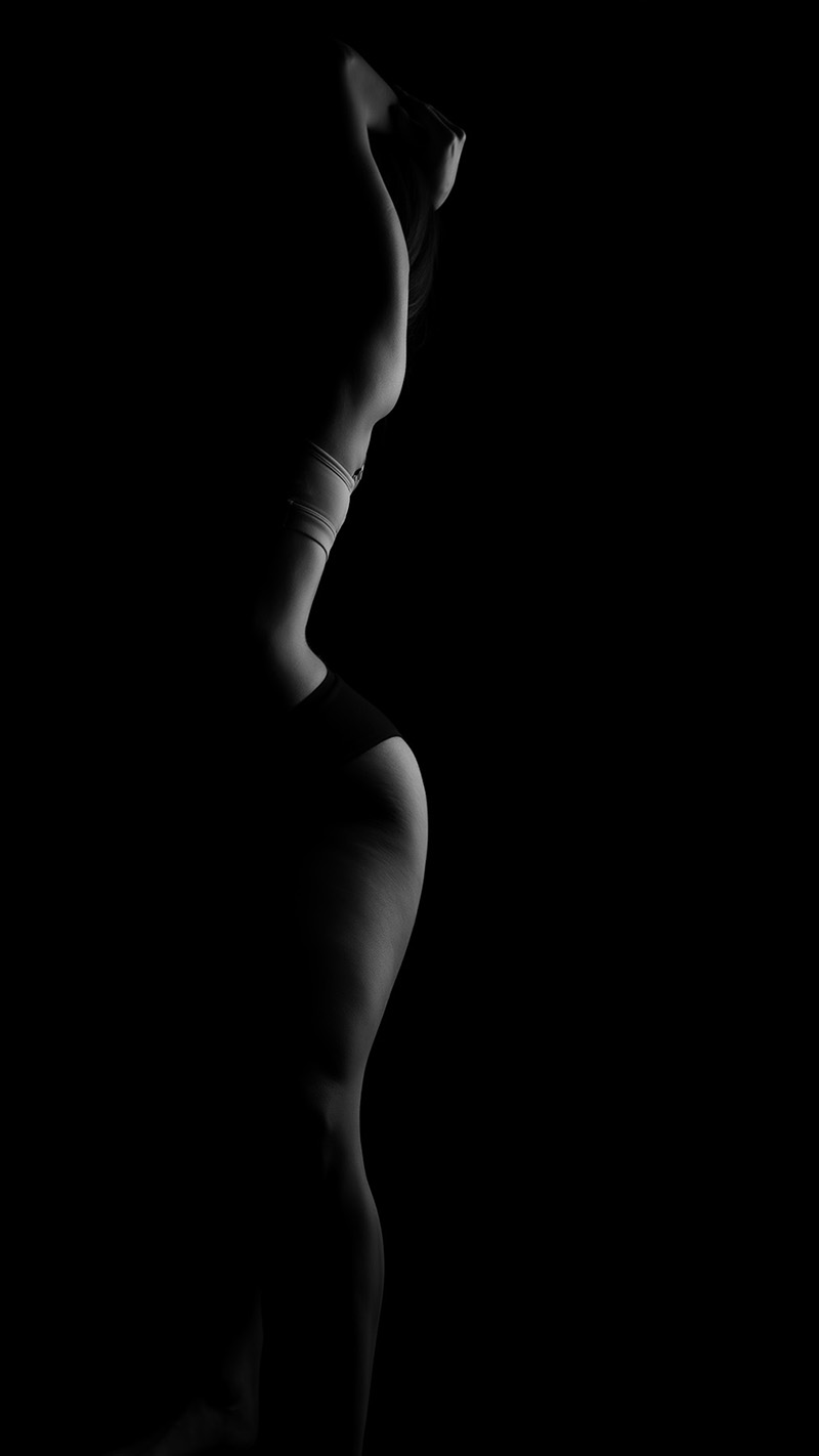 Side view of a person in minimal clothing posing with arms raised, illuminated by soft light that highlights body contours against a dark background