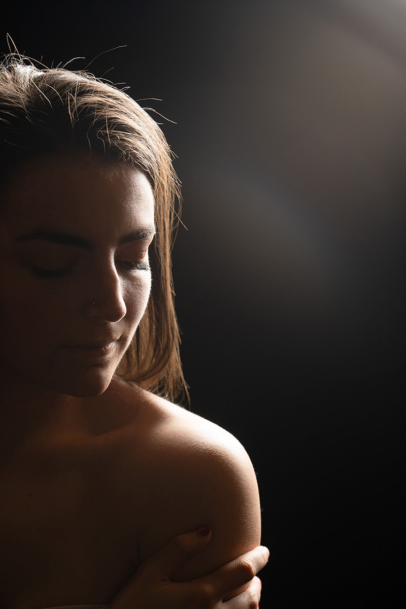 Young woman with closed eyes gently holding her shoulder, softly illuminated against a dark background