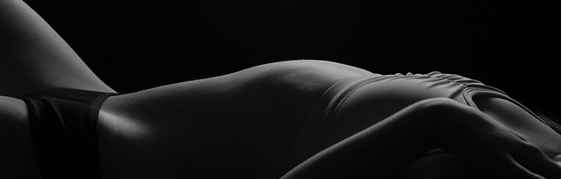 Side view of a person lying down wearing black underwear, with their torso and upper thighs highlighted by soft, low lighting against a black background