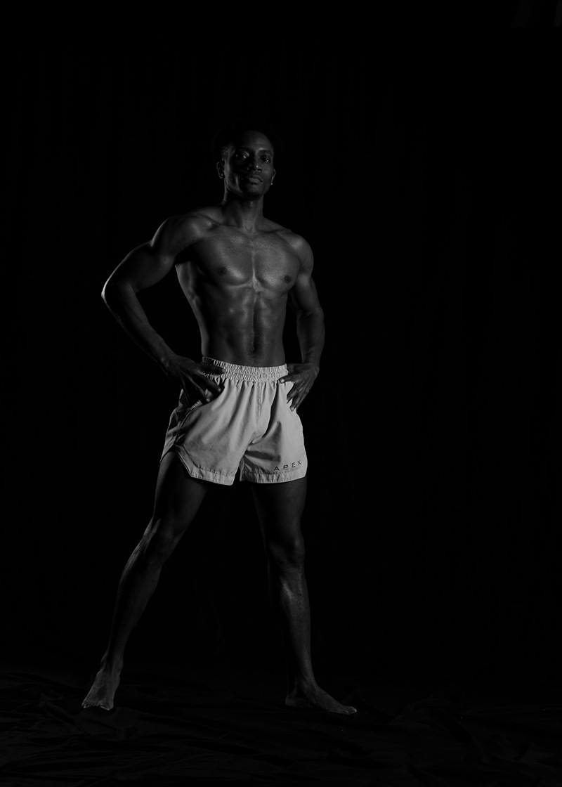 Muscular man standing barefoot in light shorts, hands on hips, against a black background