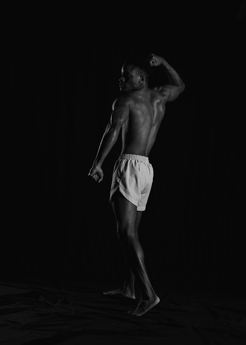 Muscular man standing barefoot against a dark background, flexing his right arm while looking over his shoulder, wearing light shorts