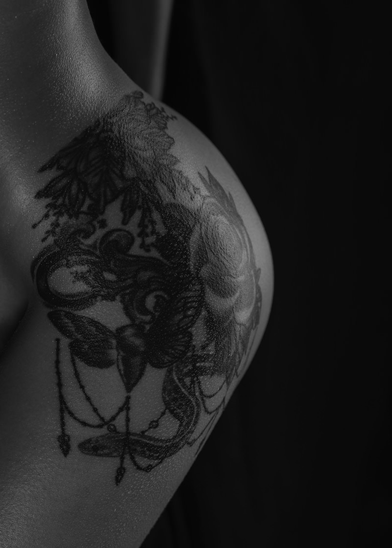 Close-up of a shoulder with a detailed black and gray tattoo featuring flowers, leaves, and hanging chains against a dark background