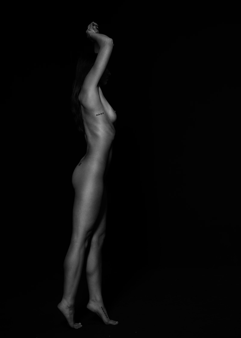 Nude woman standing on tiptoes with arms raised above head, side profile visible against a black background