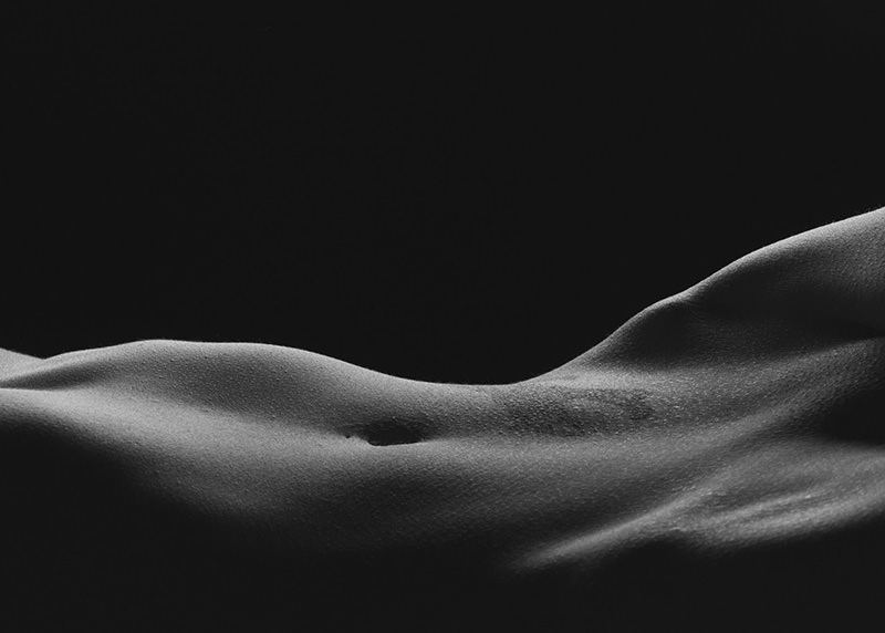 Close-up of a toned and textured upper torso with a visible navel, lit from the side against a dark background, emphasizing muscle contours and skin texture.