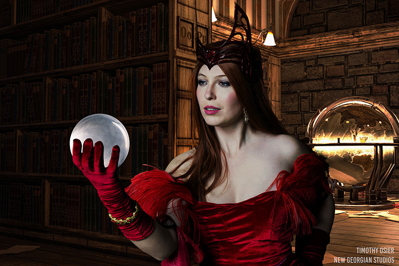Woman in a red dress and elaborate headpiece holding a glowing white orb, standing in a dimly lit room with wooden bookshelves and a large illuminated globe in the background