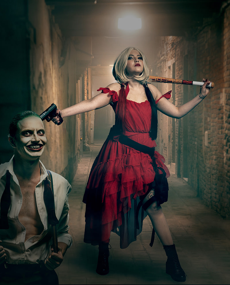 Woman dressed as Harley Quinn in a red dress with black and red ponytails holding a baseball bat over her shoulder and pointing a gun at a man dressed as Joker. The scene is set in a dimly lit brick hallway with a bright light above.
