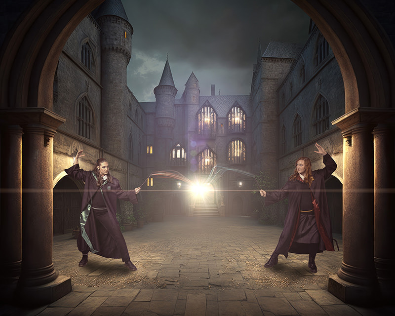 Two women in long dark robes casting magic with wands, facing each other in a stone courtyard of a castle-like building under a cloudy sky, magic light glowing between their wands