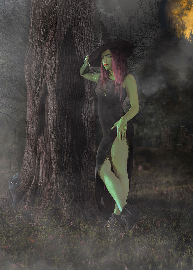 A woman dressed as a witch with a black pointed hat and black dress stands leaning against a large tree in a misty graveyard at night. Her skin is illuminated with green light, and she wears dark boots. A black cat with glowing eyes is partially visible behind the tree, and a large orange full moon shines in the dark, foggy sky.