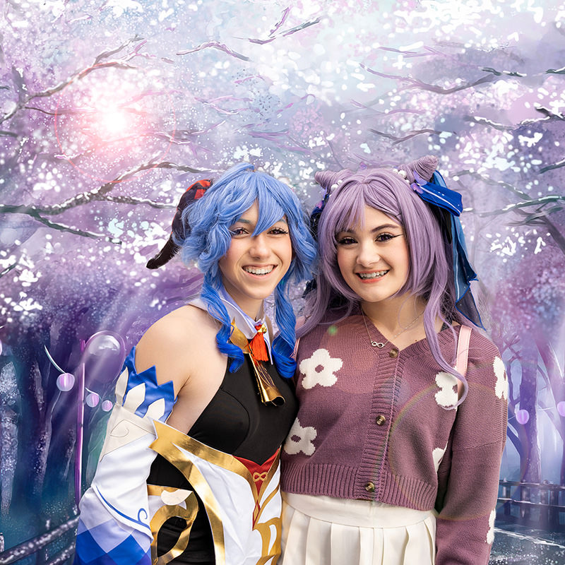 Two young women smiling and posing together, both wearing colorful cosplay costumes. The woman on the left has blue hair styled in curls and wears a black and white outfit with gold and blue accents. The woman on the right has lavender hair styled in twin buns with ribbons and wears a purple cardigan with white floral patterns and a white skirt. The background features an artistic, magical depiction of cherry blossom trees with soft glowing light effects.