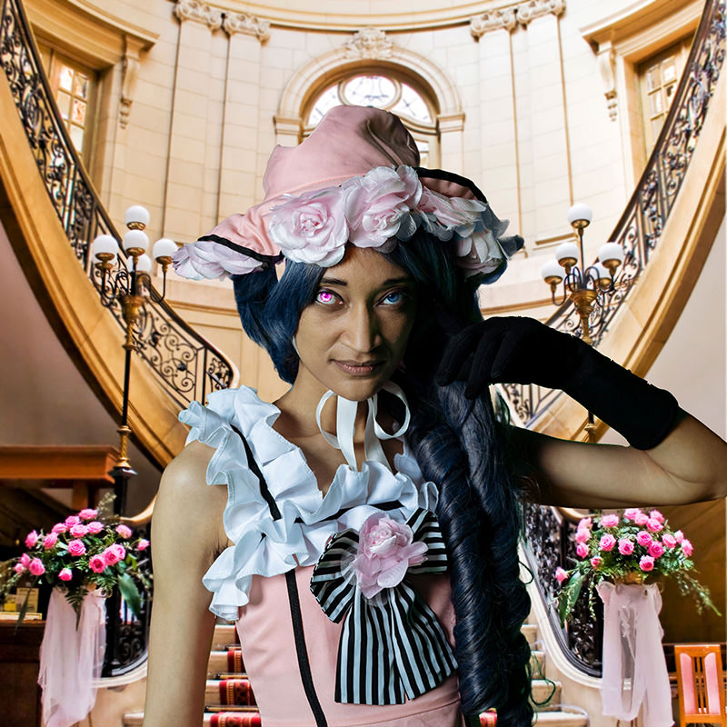 Person in a detailed cosplay costume with a large pink hat adorned with fabric flowers, long dark blue curled hair, mismatched colored eyes, wearing a pink dress with ruffles and striped bow, posing inside a grand staircase with floral arrangements and ornate railings