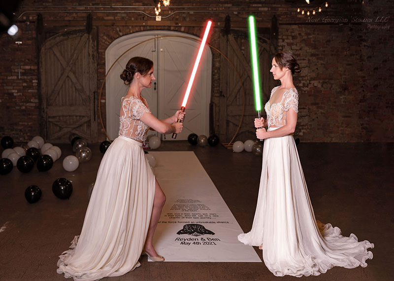 Two brides in elegant white dresses holding glowing lightsabers, one red and one green, facing each other in a rustic venue decorated with scattered black, white, and silver balloons and a white aisle runner with wedding details.