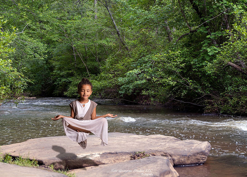 A young girl with closed eyes and calm expression sits cross-legged in a meditation pose, levitating above a large flat rock near a flowing river surrounded by dense green trees.