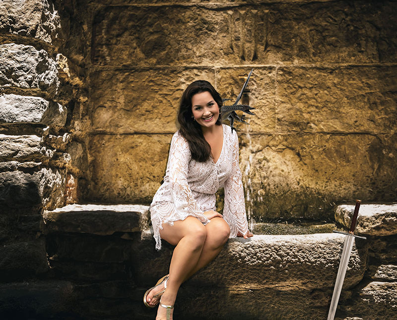A woman wearing a white lace dress sits on a stone ledge in front of a textured stone wall. A small, black, winged dragon rests on her shoulder, and a medieval-style sword with a brown leather-wrapped handle leans against the stone ledge beside her.