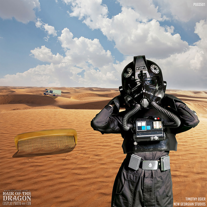 Person wearing a black sci-fi pilot helmet and suit stands in a sandy desert with rolling dunes under a cloudy blue sky, a large brown comb floats in the air nearby, and a vehicle is faintly visible in the distance on the sand.