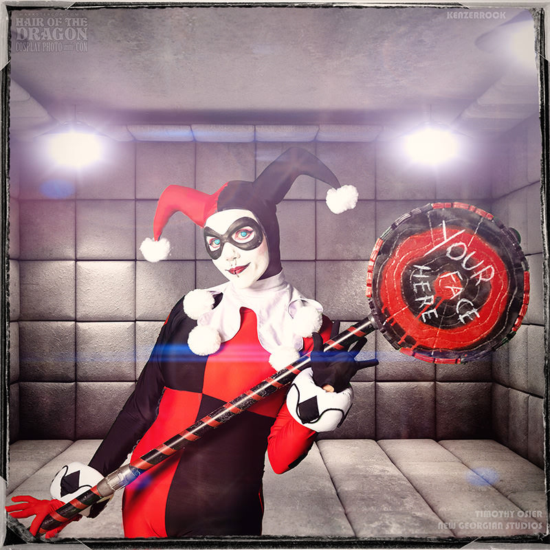 Person dressed as Harley Quinn in a red and black jester costume holding a large red and black hammer with 'Your Face Here' written on it, standing in a padded gray room.