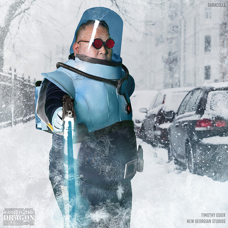 Person wearing a blue futuristic armored suit with a transparent helmet and red goggles, holding a glowing weapon, standing in a snowy city street with parked cars covered in snow